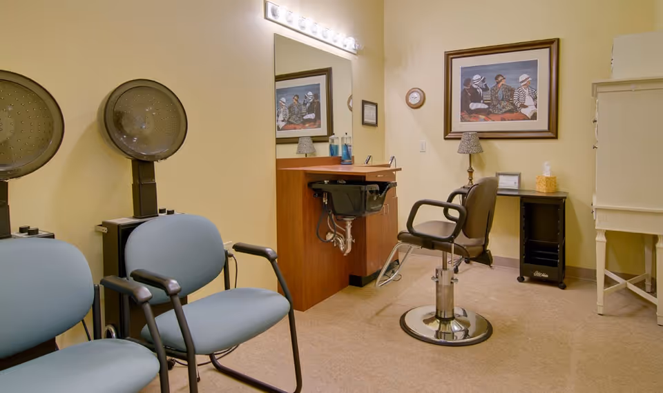 Interior of a senior living facility hair salon with two blue waiting chairs, two hair dryer chairs, a styling chair in front of a sink and mirror, a small table with a lamp and tissue box, and a framed picture on the wall.