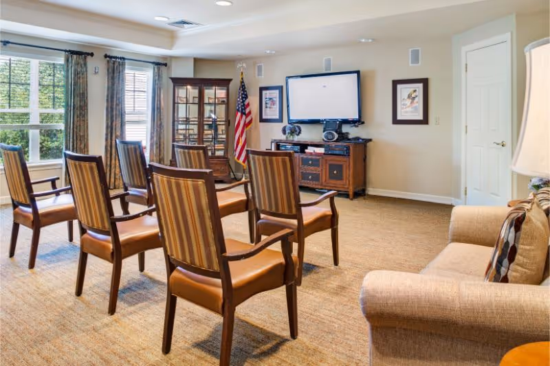 A well-lit common room with six wooden chairs arranged in two rows facing a flat-screen TV mounted on the wall. The room features beige walls, a carpeted floor, a beige sofa with patterned pillows, an American flag, framed artwork, a glass display cabinet, and large windows with curtains allowing natural light to enter.