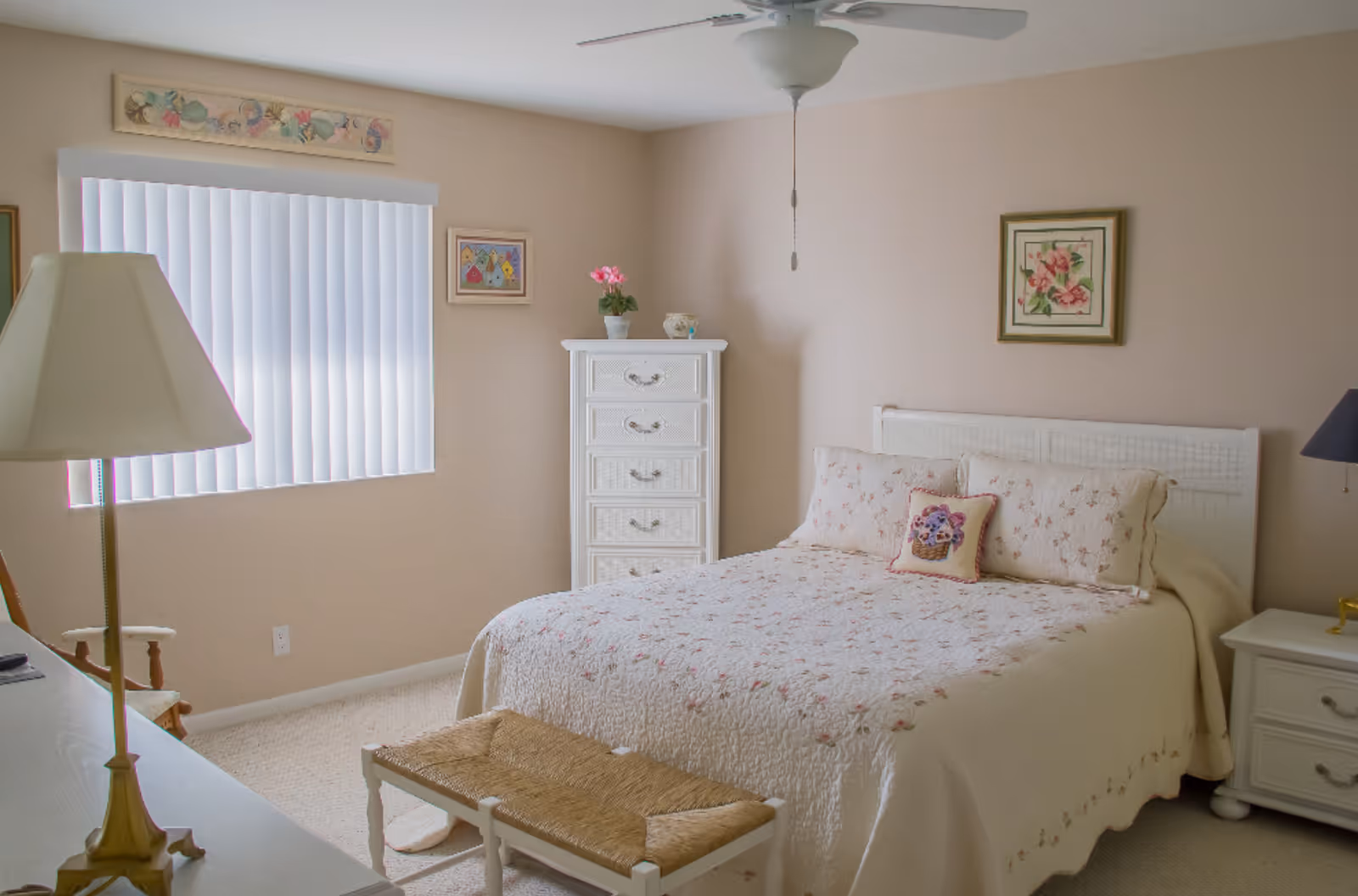 A cozy bedroom with a large bed covered in a white quilt with floral patterns. There are matching pillows on the bed, including a decorative pillow with a flower basket design. A white dresser with five drawers stands against the wall, topped with a small potted plant and a decorative item. A white nightstand with a lamp is beside the bed. The room has beige walls adorned with framed floral artwork, a window with vertical blinds, a ceiling fan, and a woven bench at the foot of the bed.