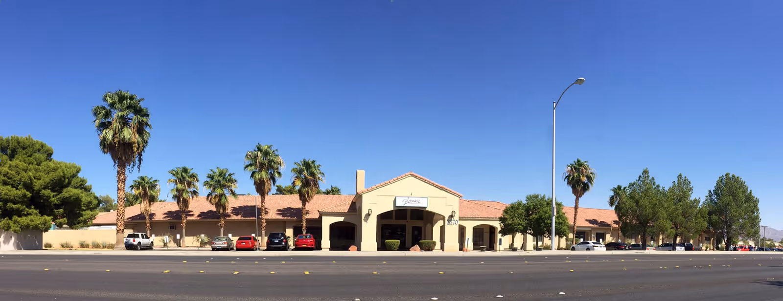 Front exterior of a single-story Mediterranean-style senior living facility with a tile roof, palm trees, and cars parked along a wide street.