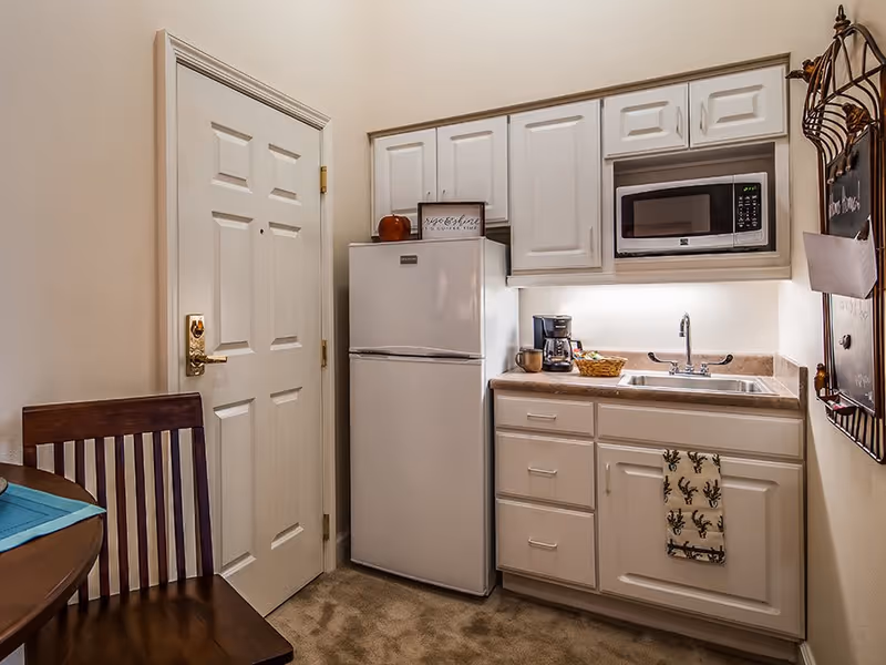 Small kitchen area with white cabinets, a white refrigerator, a microwave, a coffee maker on the countertop, and a sink. There is a wooden chair and part of a round table with a blue placemat visible on the left side. A door with a gold handle is next to the refrigerator.