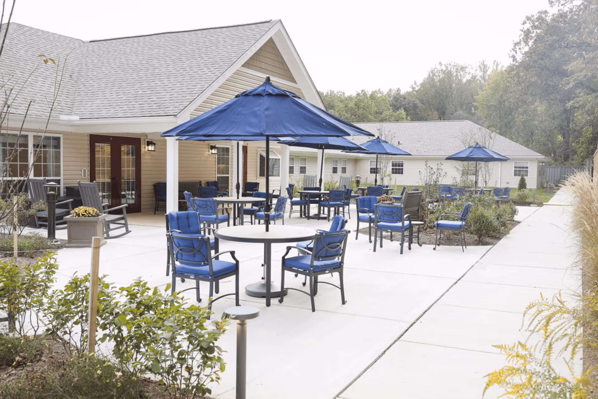 Outdoor patio area at a senior living facility with multiple round tables and blue cushioned chairs under blue umbrellas. The patio is surrounded by plants and shrubs, with a beige building in the background and a concrete walkway on the right side.
