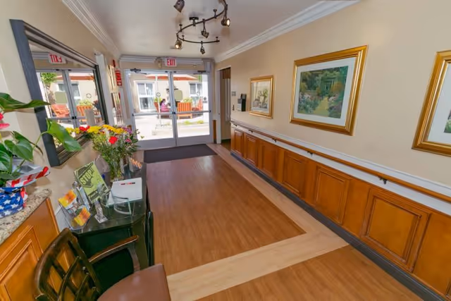 Interior hallway of Arroyo Vista Nursing Center with wooden flooring, handrails along the wall, framed paintings, a small table with flowers and brochures, and glass double doors leading outside.