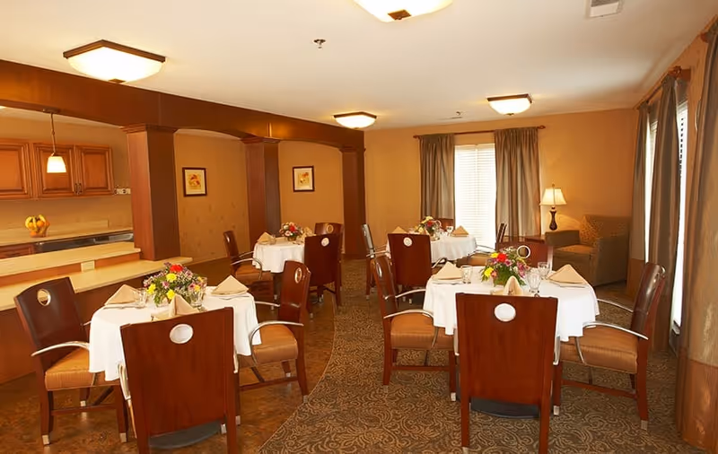 Small dining room with round tables set with white tablecloths, floral centerpieces, wooden chairs, and a seating area by the windows.