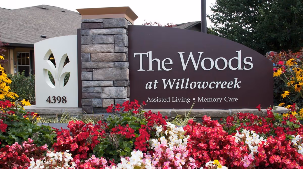 Entrance sign for The Woods at Willowcreek Assisted Living and Memory Care mounted by a stone pillar and surrounded by colorful flowers.
