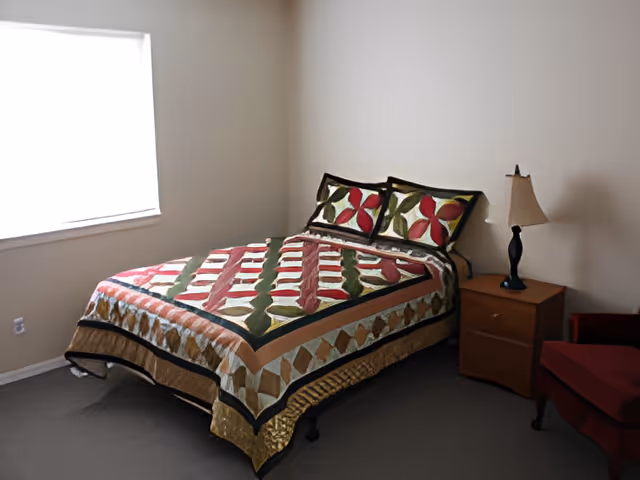 A neatly made bed with a colorful patterned quilt, a nightstand with a lamp, a chair, and a window in a simple bedroom.