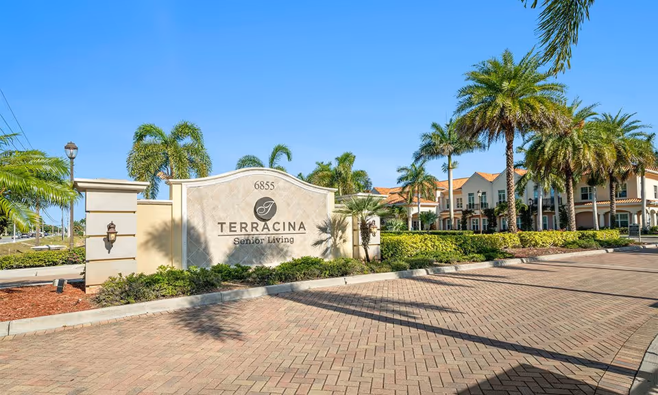 Entrance sign for Terracina Senior Living with the address 6855, surrounded by palm trees and landscaping, with the senior living facility building visible in the background under a clear blue sky.