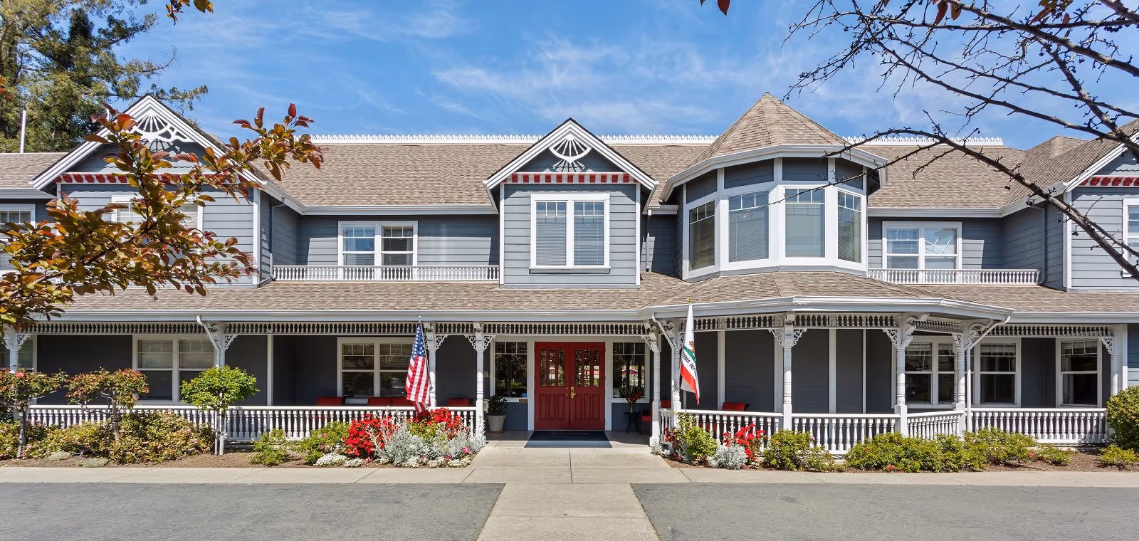 Front exterior view of a large, two-story senior living facility building with gray siding, white trim, and a covered porch. The entrance features double red doors, American and California state flags, and landscaped flower beds with shrubs and red flowers. The sky is clear and blue.