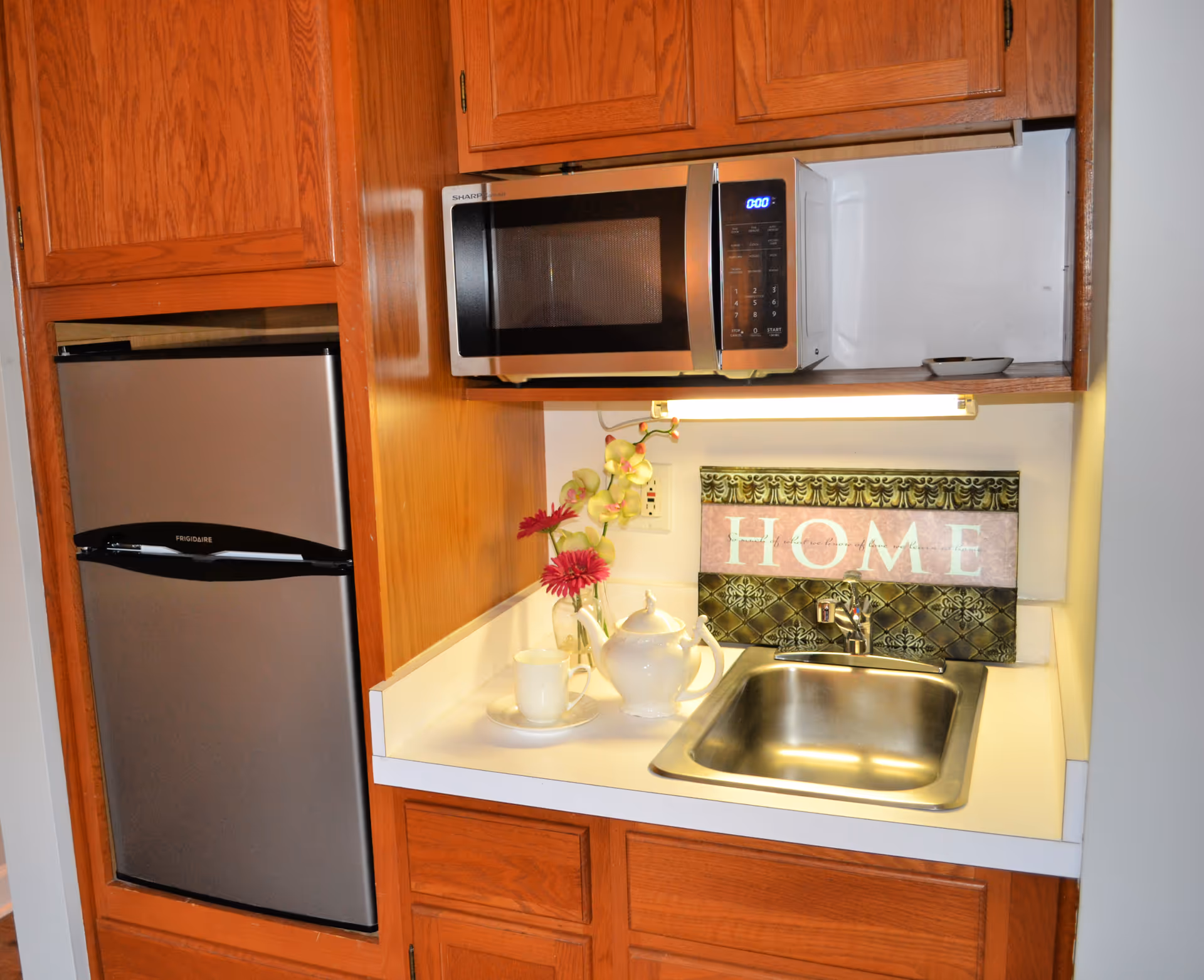 Small kitchenette with a stainless steel mini-fridge, microwave, sink, teapot and flowers, and a decorative sign reading 'HOME'.