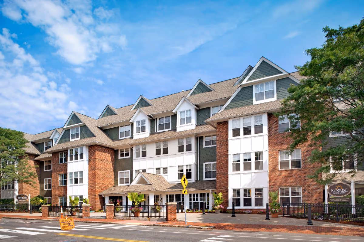 Exterior view of a multi-story senior living facility building with a brick and white facade, multiple windows, and a pitched roof under a partly cloudy blue sky. There is a sidewalk, a pedestrian crossing sign, and a detour sign in front of the building. Trees and landscaping are visible around the entrance.