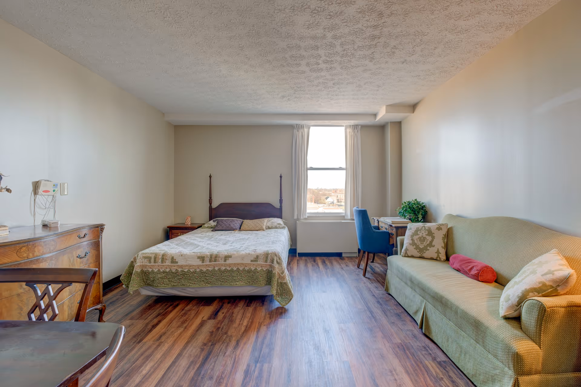 A senior living facility bedroom with a bed covered in a patterned quilt, a wooden dresser, a small wooden nightstand, a green sofa with decorative pillows, a blue upholstered chair, and a wooden desk near a window with white curtains. The room has wood flooring and beige walls.