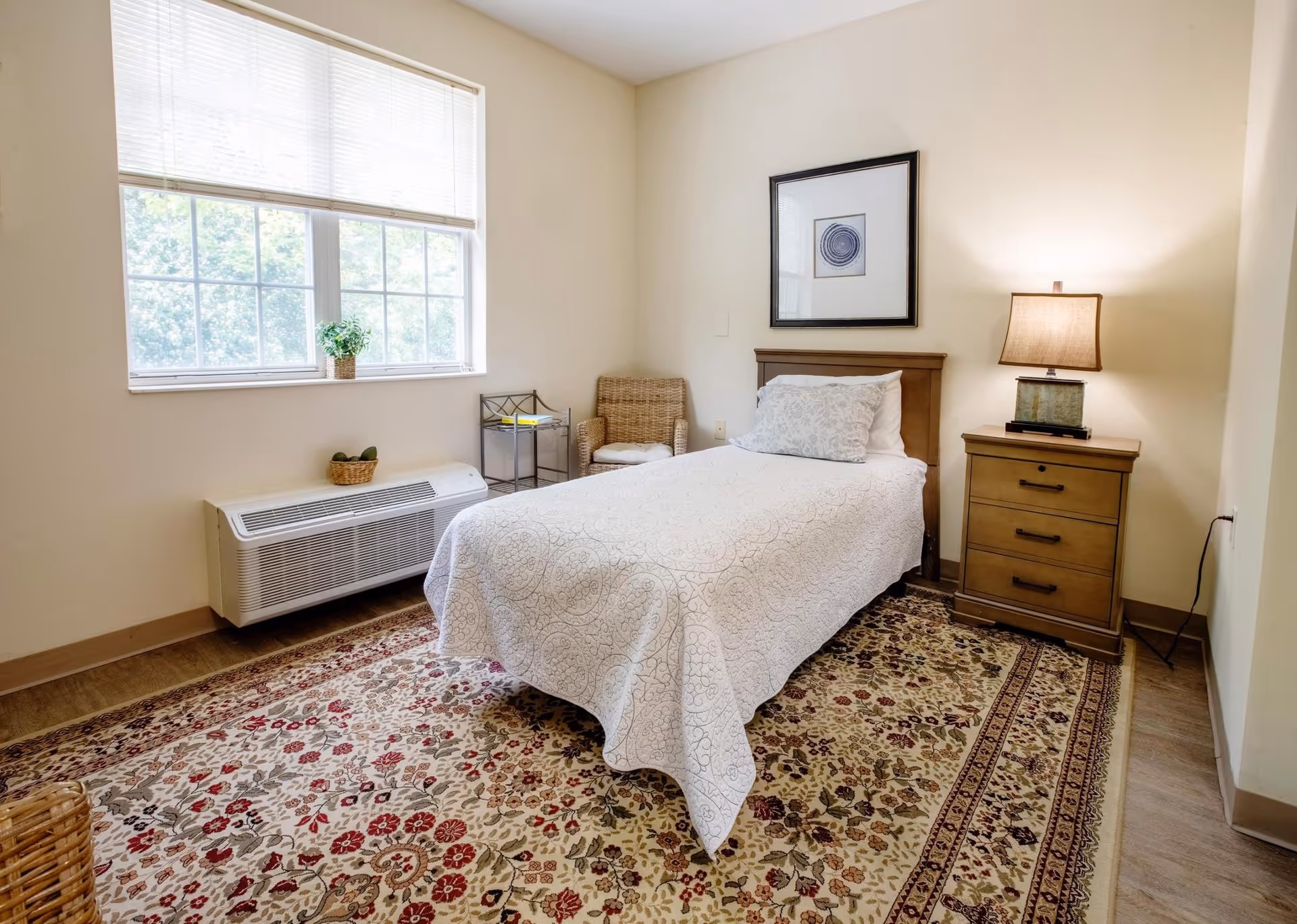 Simple single-occupancy bedroom with a twin bed, wooden nightstand and lamp, window with blinds, wicker chair, and patterned area rug.