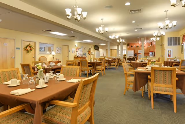 A spacious dining room with multiple tables covered in brown tablecloths, each set with white cups, glasses, and napkins. The room is well-lit with chandeliers hanging from the ceiling and has wooden chairs with patterned upholstery. The walls are painted light yellow, and there is a kitchen area visible in the background.