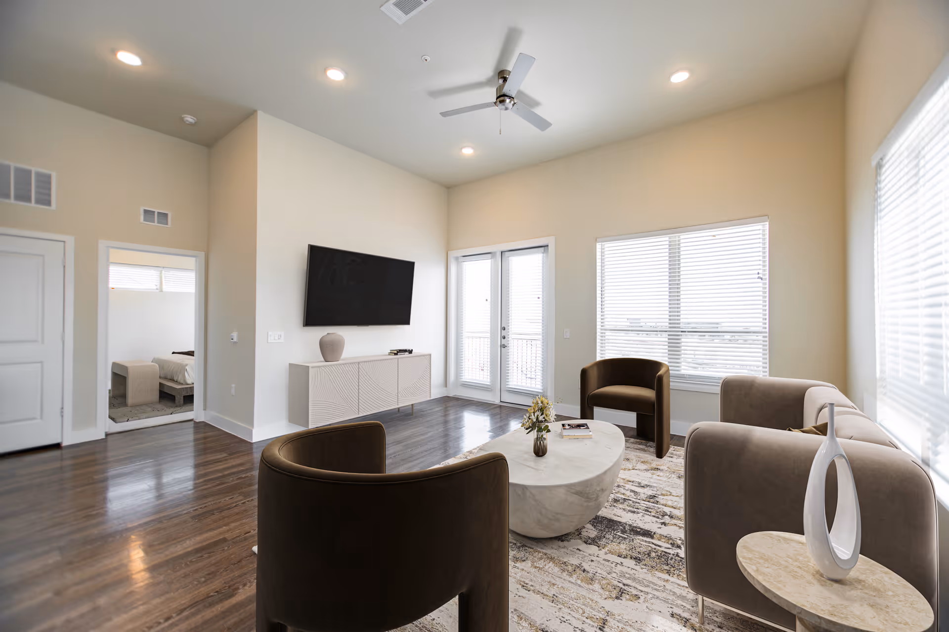 A bright and modern living room with wooden flooring, a ceiling fan, a wall-mounted flat-screen TV above a white cabinet, two brown armchairs, a beige sofa, a marble coffee table with a small flower vase, and large windows with blinds allowing natural light. A doorway leads to a bedroom with a bed visible.