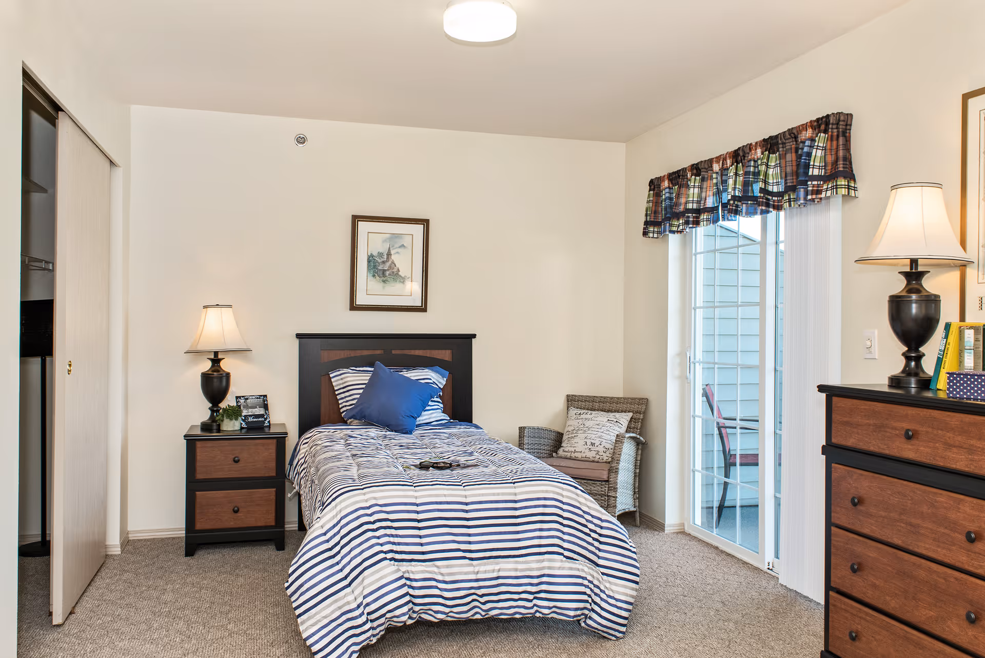 A cozy bedroom with a single bed covered in a striped blue and white comforter and a blue pillow. There is a nightstand with a lamp and small plant to the left of the bed, and a wicker chair with a cushion to the right near a sliding glass door with plaid valance curtains. A dresser with a lamp and books is on the right side of the room, and a framed picture hangs on the wall above the bed.
