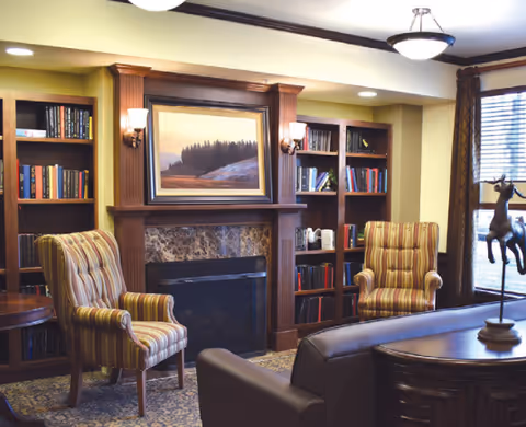 A cozy living room area featuring a fireplace with a landscape painting above it, flanked by built-in bookshelves filled with books. Two striped armchairs are positioned near the fireplace, and a dark leather sofa is partially visible in the foreground. The room is warmly lit with wall sconces and ceiling lights, and a window with brown curtains allows natural light to enter.