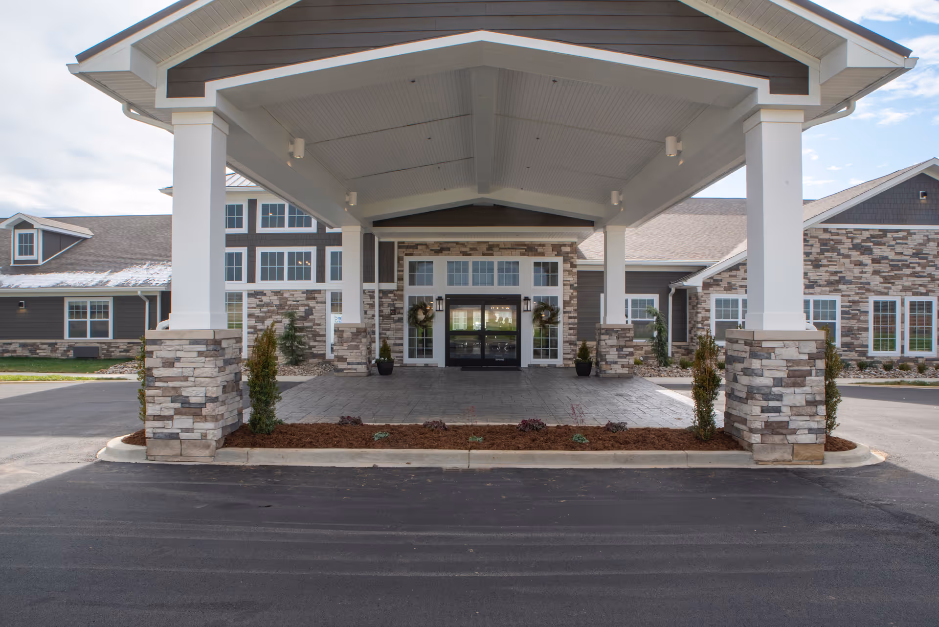 Entrance of a senior living facility with a covered driveway supported by white pillars with stone bases, leading to double glass doors decorated with wreaths. The building exterior features a combination of stone and dark siding with multiple windows.