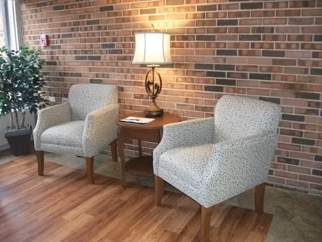 Two upholstered armchairs flanking a wooden side table with a lamp against an exposed brick wall.