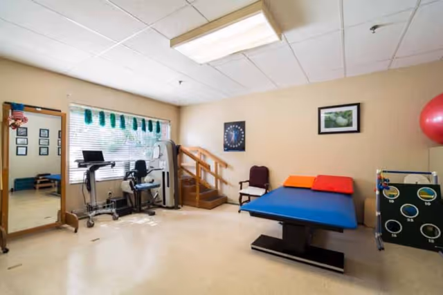 A physical therapy or rehabilitation room with exercise equipment, a large mirror, a treatment table with colorful cushions, a chair, and a small staircase with handrails. The room has beige walls, a window with blinds, and framed pictures on the wall.