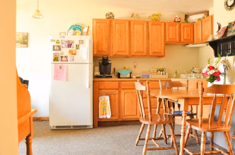 A cozy kitchen and dining area featuring wooden cabinets, a white refrigerator with magnets and notes, a coffee maker on the counter, and a wooden dining table with four matching chairs. There are decorative items on top of the cabinets and a vase with flowers on the table.