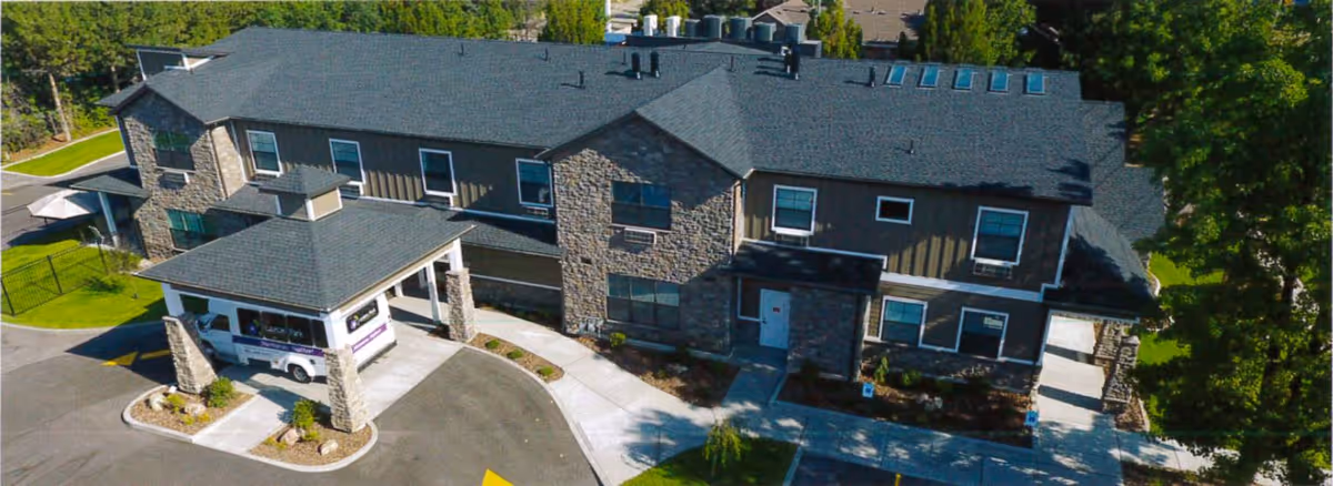 Aerial view of a two-story senior living facility building with stone and siding exterior, a covered entrance with a vehicle parked underneath, surrounded by trees and landscaping.