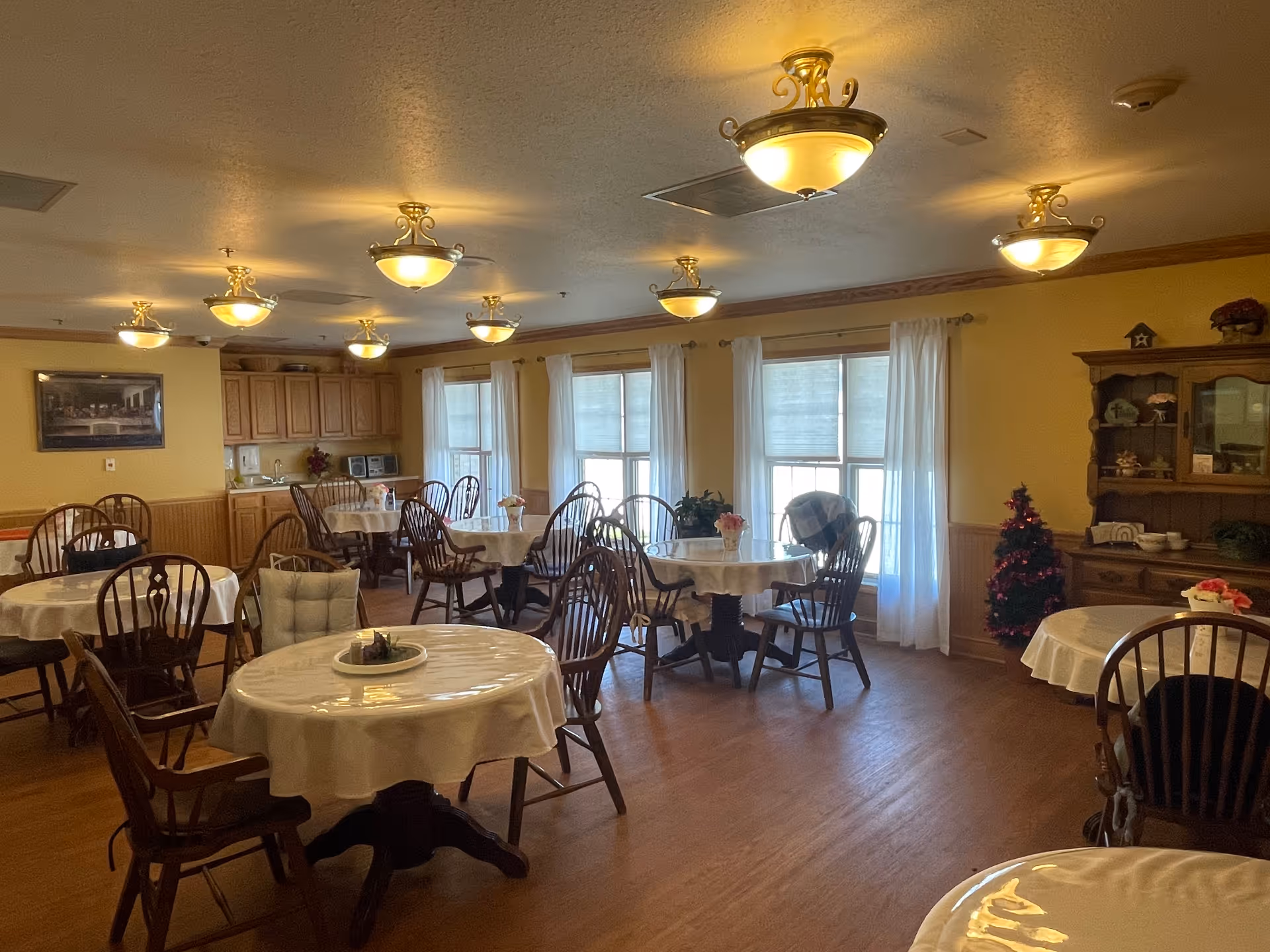 A dining room in Serenity Assisted Living with several round tables covered with white tablecloths and surrounded by wooden chairs. The room has large windows with white curtains allowing natural light to enter. There are multiple ceiling lights providing warm illumination. A wooden cabinet with decorative items and a small Christmas tree are visible in the corner. The walls are painted yellow with wood paneling on the lower half.