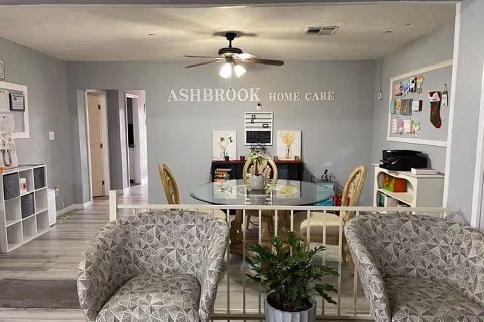 Interior view of a senior living facility room with two patterned armchairs in the foreground, a small green plant in a pot, and a glass dining table with four chairs in the background. The wall behind the table has the text 'ASHBROOK HOME CARE' displayed. The room has light gray walls, a ceiling fan with lights, and various decorations including framed pictures and a bulletin board.