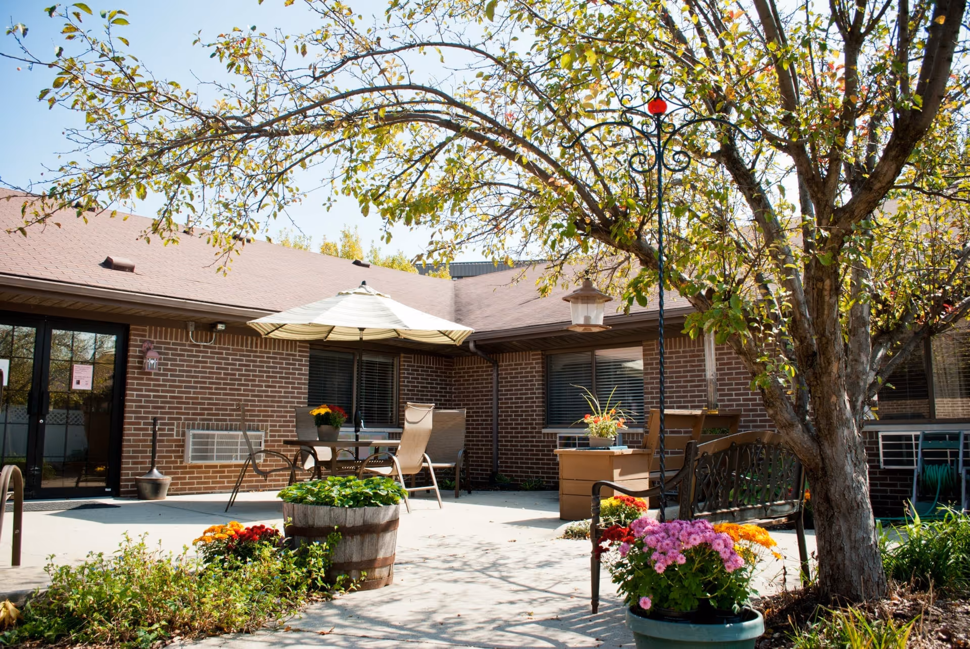 Outdoor patio area at Bethany Village with a large tree, potted flowers, a metal bench, a table with chairs under a large umbrella, and a brick building in the background.
