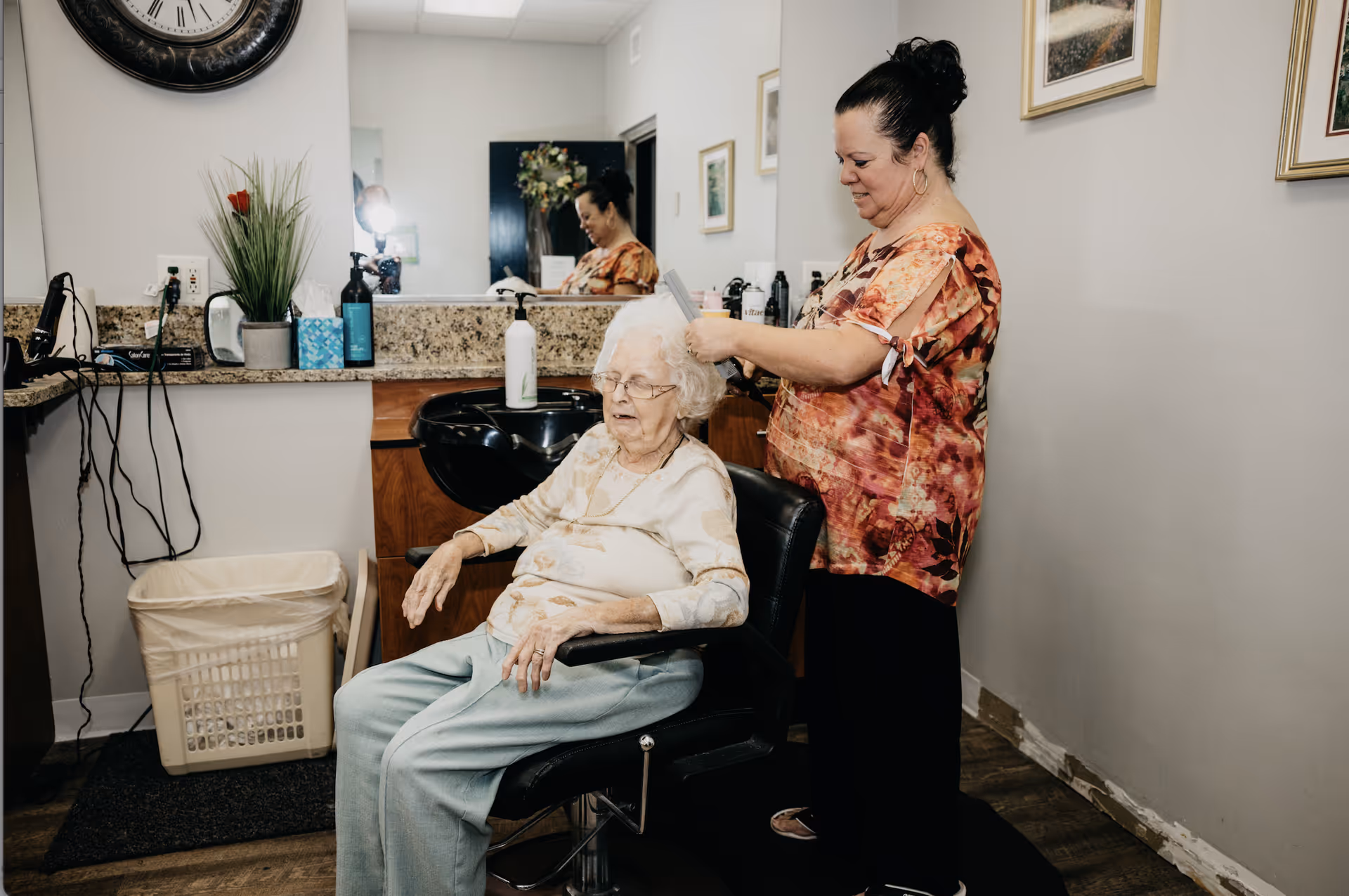 An elderly woman with white hair sits in a salon chair while a hairstylist stands behind her, combing her hair. The room has a large mirror, a countertop with hair care products, a clock on the wall, and framed pictures. A laundry basket is visible on the floor.