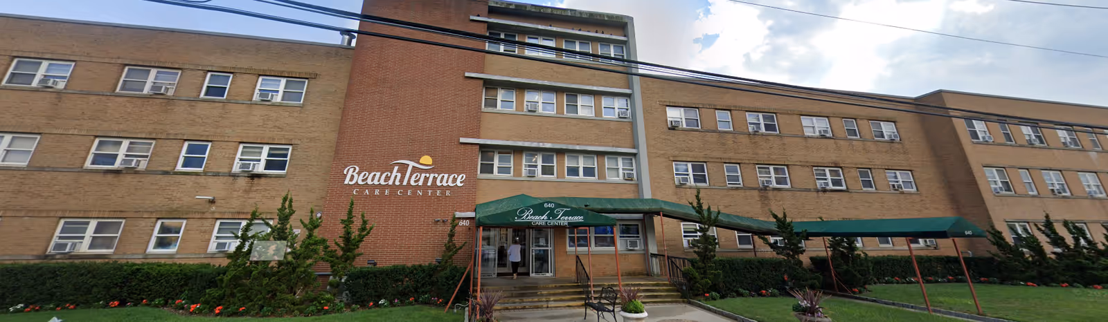 Front exterior view of the Beach Terrace Care Center, a multi-story brick building with numerous windows. There is a green awning over the entrance with the facility name on it, and a walkway leading up to the entrance. The surrounding area has green grass, bushes, and some small trees.