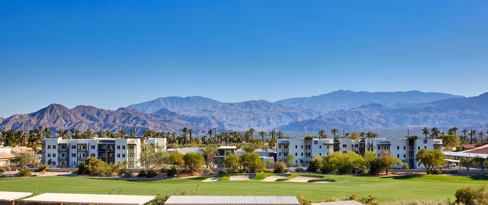 Wide exterior view of Revel Palm Desert facility with modern white buildings surrounded by palm trees and greenery, set against a backdrop of rugged mountains under a clear blue sky.
