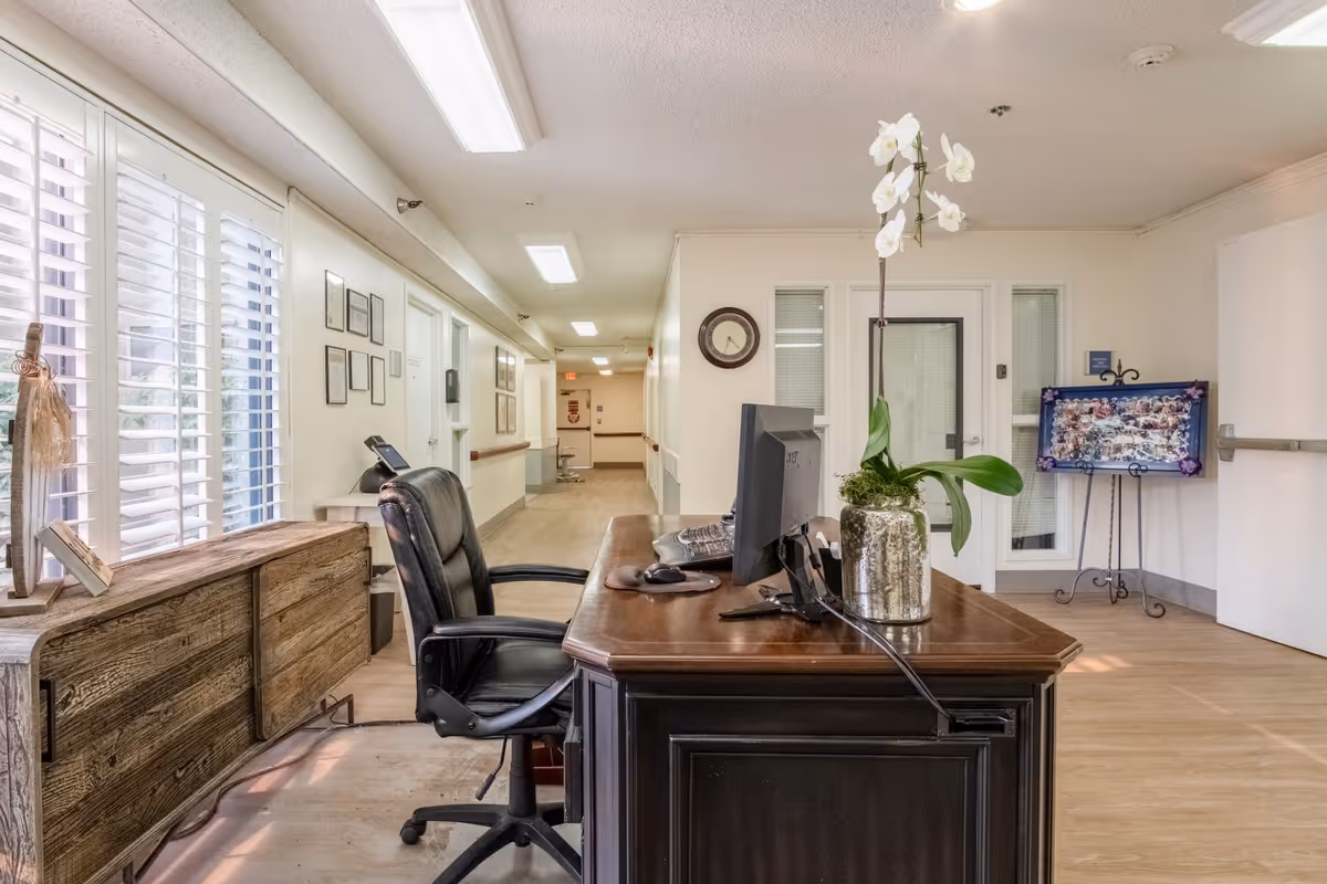 Reception area inside Vista Blue Mountain Assisted Living & Memory Care featuring a wooden desk with a computer monitor and a potted orchid plant. There is a black office chair behind the desk, a wooden sideboard with decorative items near a window with white shutters, and a hallway extending into the background. A clock is mounted on the wall, and a colorful framed artwork is displayed on an easel near a door.