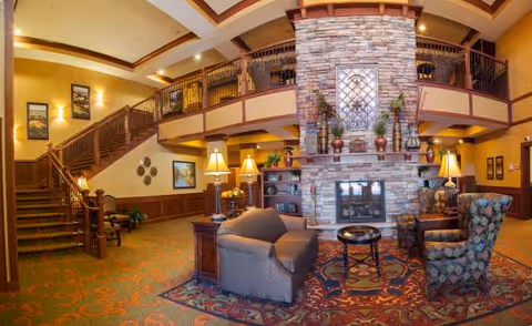 Cozy senior living lobby with a stone fireplace, seating area, staircase, and upper balcony.