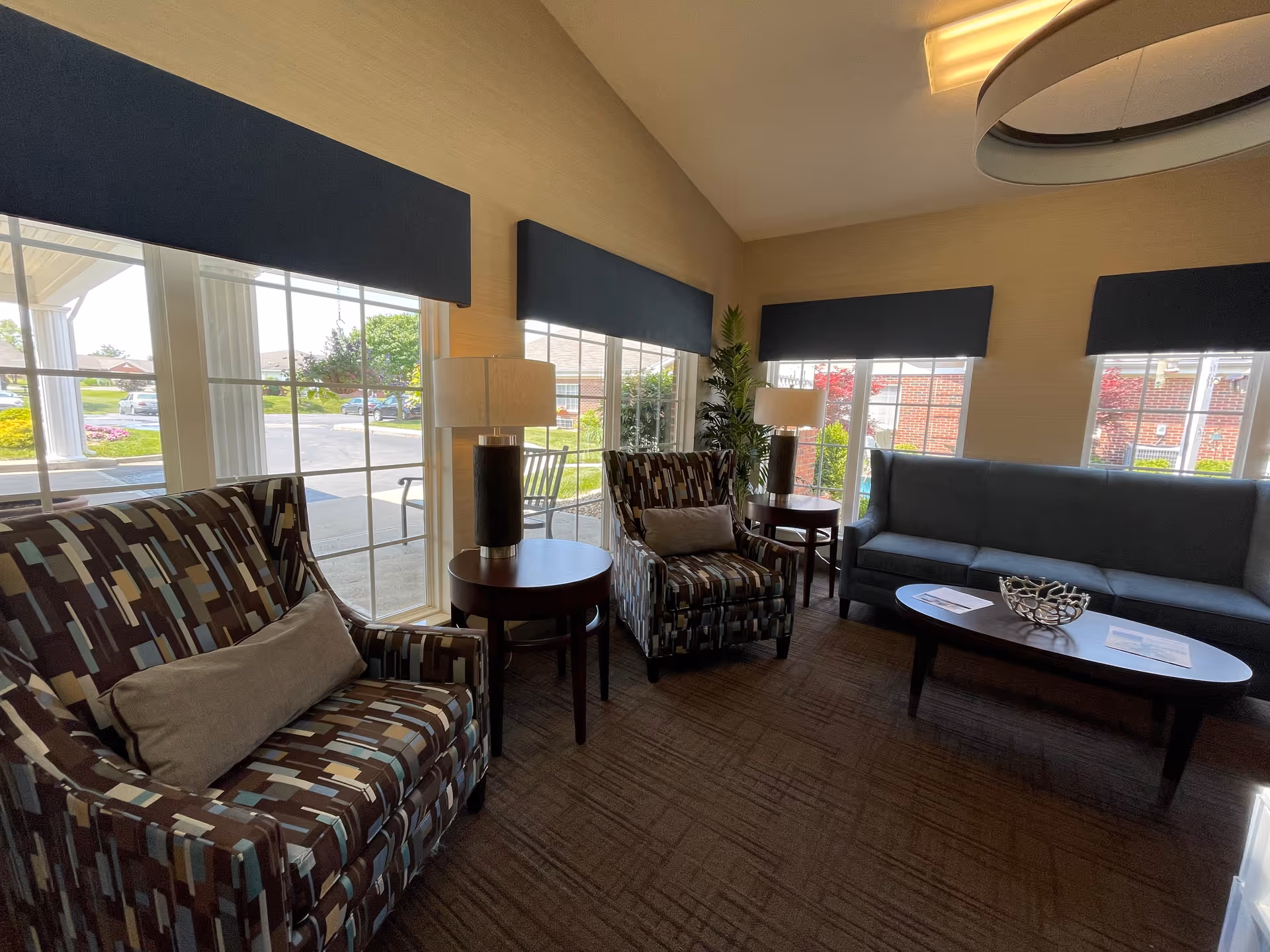 Well-lit seating area with patterned armchairs, a sofa, side tables and large windows in a retirement community lobby.