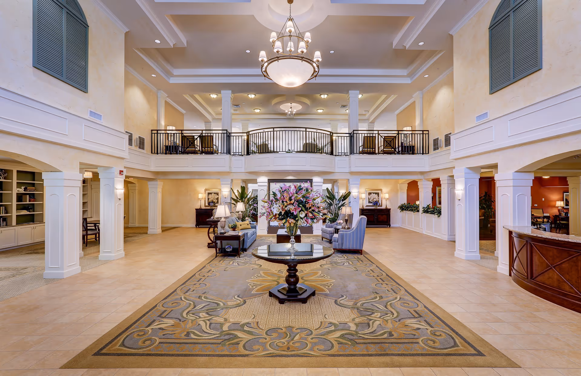 Spacious and elegant senior living facility lobby with a large decorative rug, a round table with a floral arrangement in the center, comfortable seating areas with armchairs and sofas, tall white columns, a balcony with railing on the second floor, and warm lighting from chandeliers and lamps.