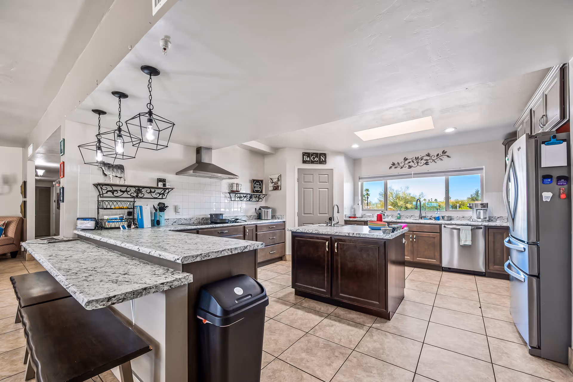 A spacious, modern kitchen with granite countertops, dark wood cabinets, and stainless steel appliances including a refrigerator and dishwasher. There is a kitchen island in the center, a breakfast bar with stools, and pendant lights hanging from the ceiling. A large window above the sink offers a view of greenery outside. The floor is tiled and the kitchen has a clean, bright appearance.