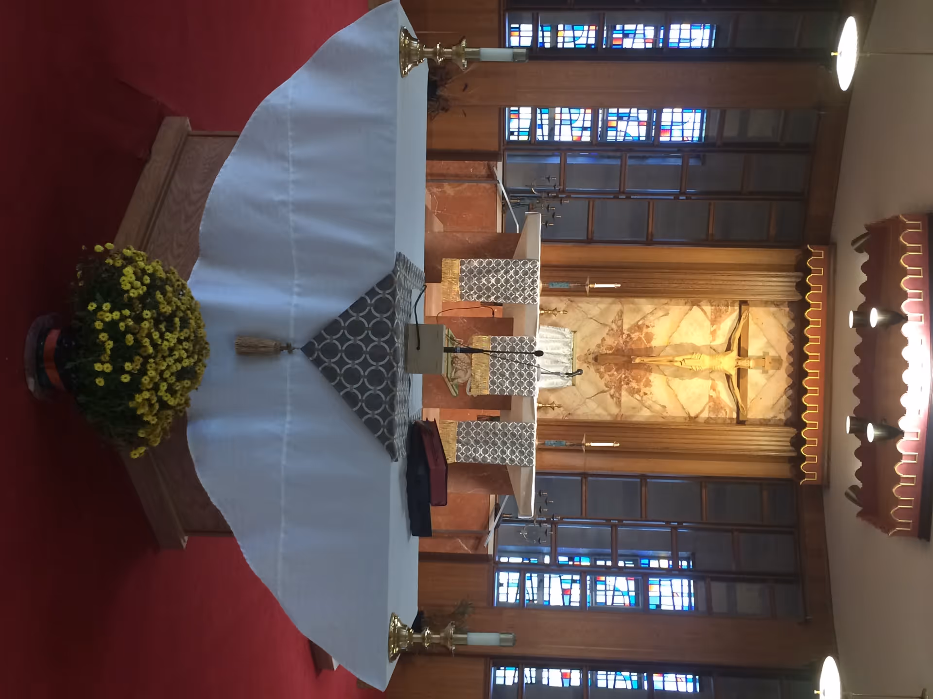 Interior view of a chapel with an altar covered by a white cloth, two candlesticks, a flower pot with yellow flowers, and a crucifix mounted on a marble wall behind the altar. Stained glass windows and wooden paneling are visible in the background.