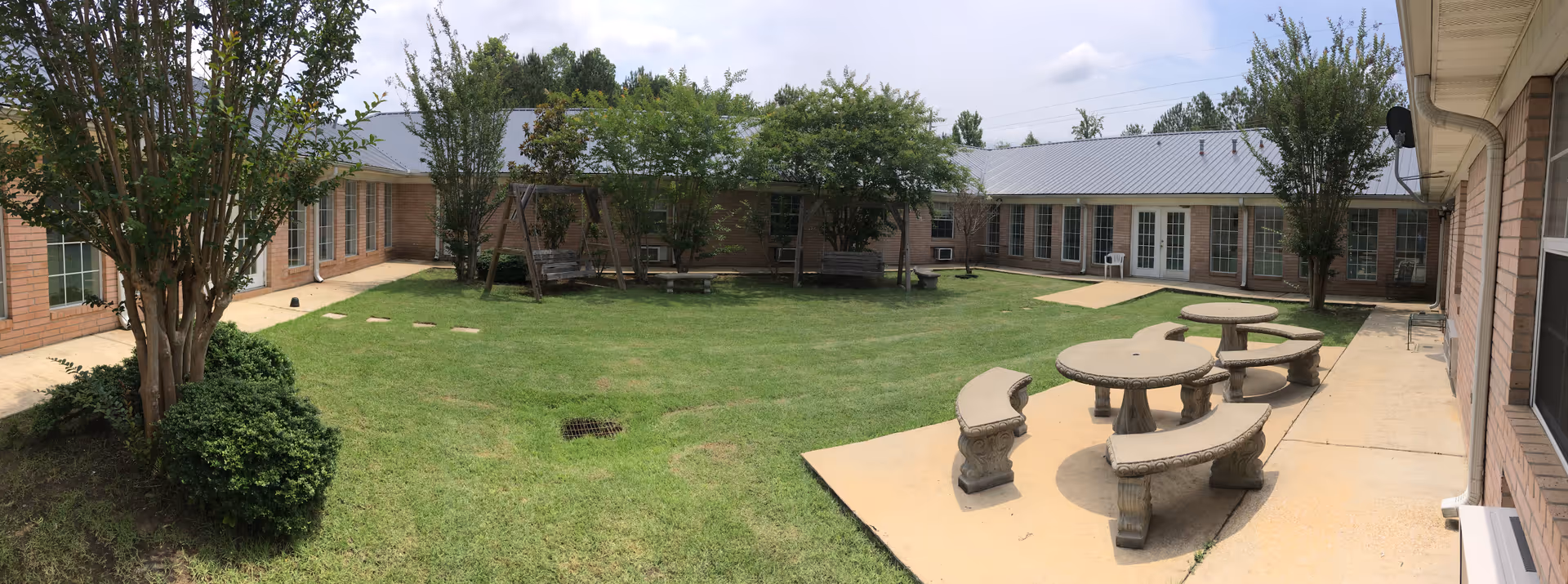 Enclosed sunny courtyard with a grassy lawn, trees, concrete picnic tables and benches surrounded by single-story brick buildings.