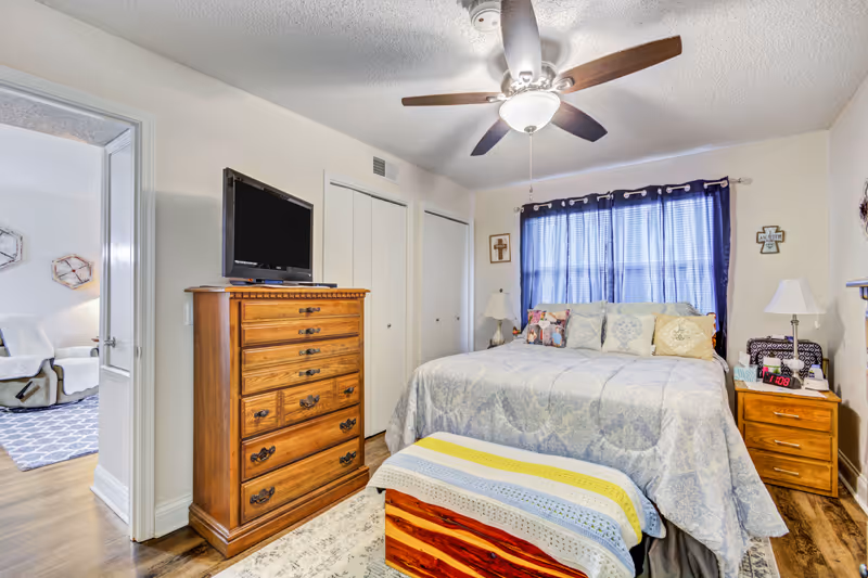 A cozy bedroom with a large bed covered in a light blue patterned comforter and multiple pillows. There is a wooden chest at the foot of the bed with a colorful knitted blanket draped over it. To the right of the bed is a wooden nightstand with a lamp, clock, and other small items. On the left side of the room is a tall wooden dresser with a flat-screen TV on top. The room has a ceiling fan with lights, blue curtains covering the window, and two closed white closet doors. The doorway on the left leads to another room with a chair and a rug visible.