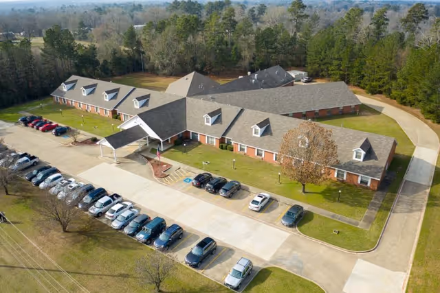 Aerial view of Legend Oaks Healthcare And Rehabilitation Center - Gladewater, showing a large single-story building with a gray roof and multiple dormer windows. The building is surrounded by a parking lot with several cars parked, green lawns, and trees in the background.