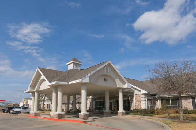 Exterior view of Legend Oaks Healthcare and Rehabilitation Center - Northwest Houston showing the main entrance with a covered drop-off area supported by white columns, a brick and beige facade, a clear blue sky with some clouds, and a few parked cars to the left.