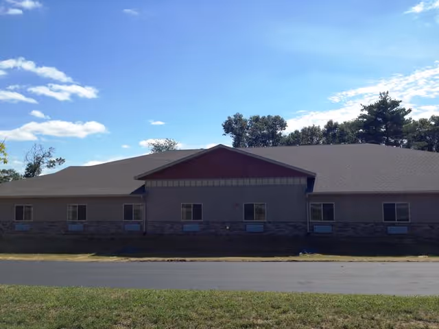 Exterior view of a single-story building with a sloped roof, multiple windows, and a stone base. The building is set against a backdrop of trees and a partly cloudy blue sky.