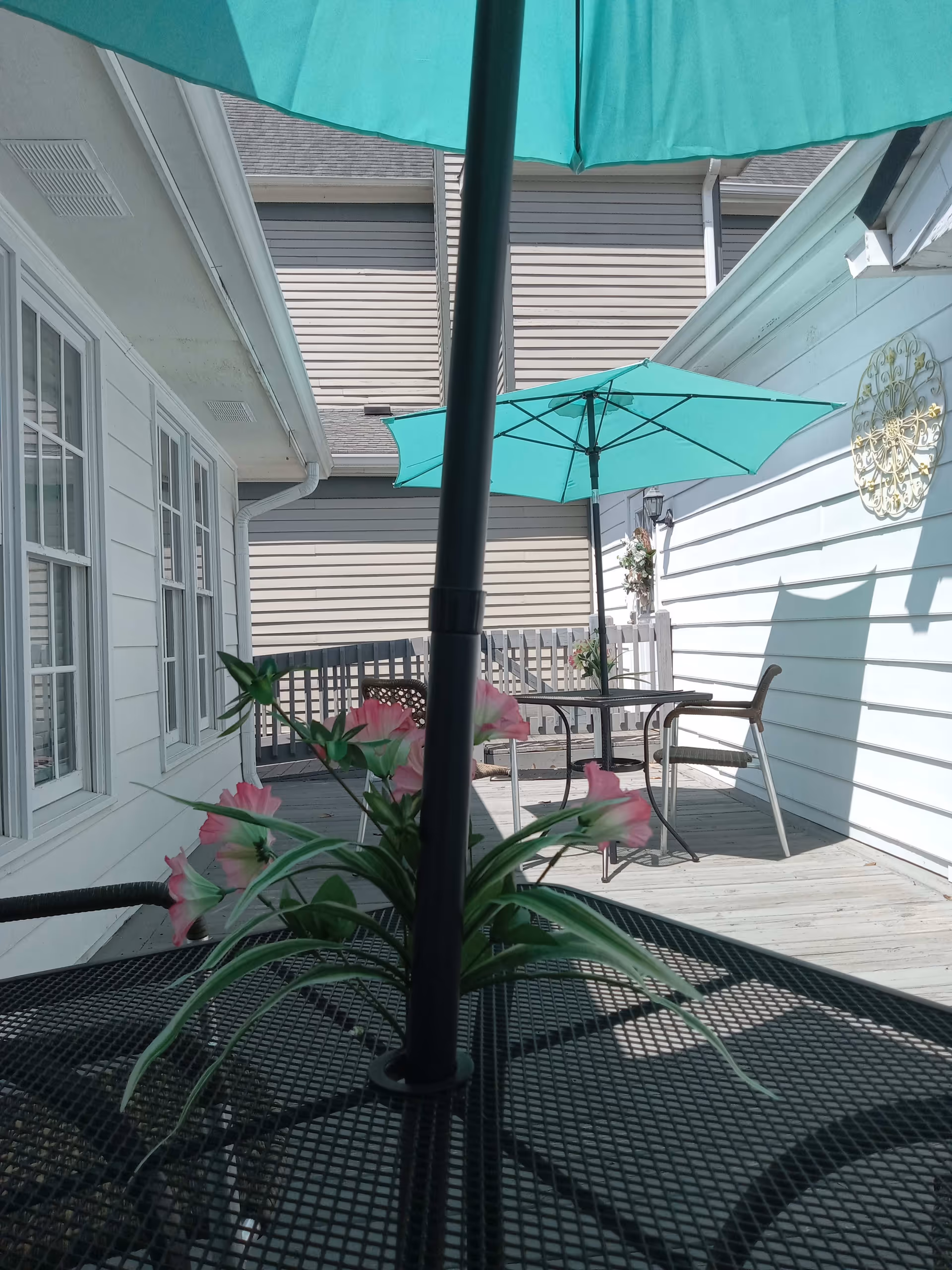 Outdoor patio area with black metal tables and turquoise umbrellas. Pink flowers are placed in a vase on the table in the foreground. The patio is surrounded by white siding walls and has a decorative golden wall hanging on one side.