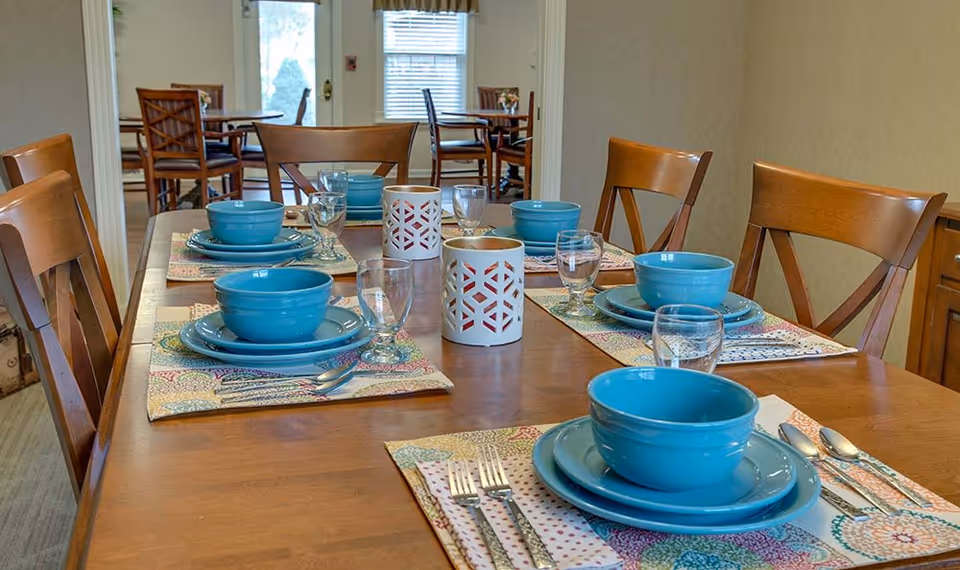 A dining table set with turquoise dishes, placemats, glassware, and wooden chairs in a communal dining room.