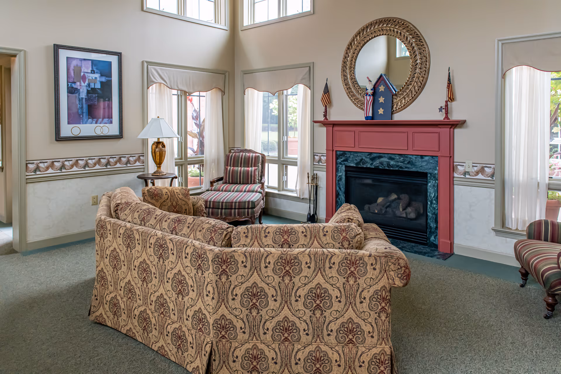 A cozy senior living community lounge area with a patterned sofa and matching armchair, a red fireplace with a decorative round mirror above it, and windows with sheer curtains letting in natural light. There is a side table with a lamp and framed artwork on the wall.