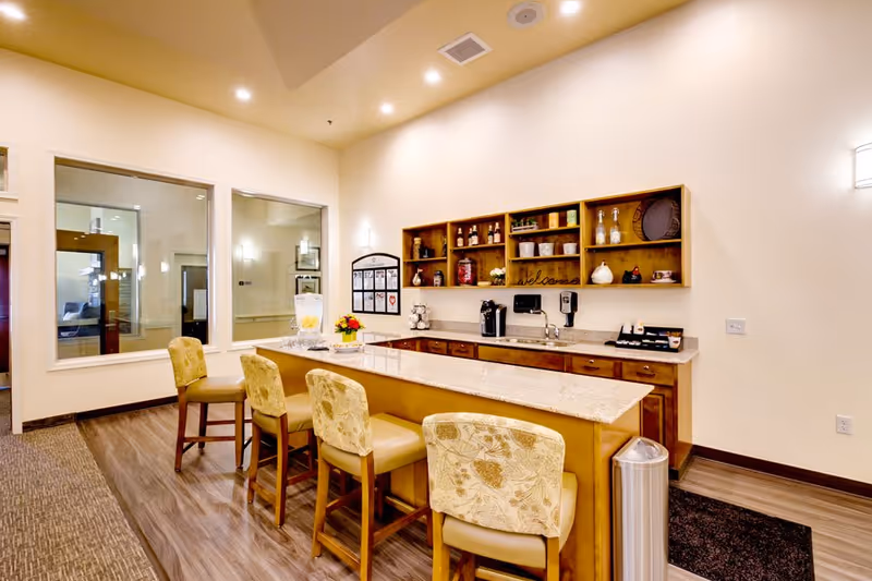 Interior view of a senior living facility's kitchen area with a long island countertop surrounded by four cushioned chairs. The back wall features wooden cabinets and shelves with various decorative items and kitchen supplies. The room has warm lighting and large windows looking into another room.