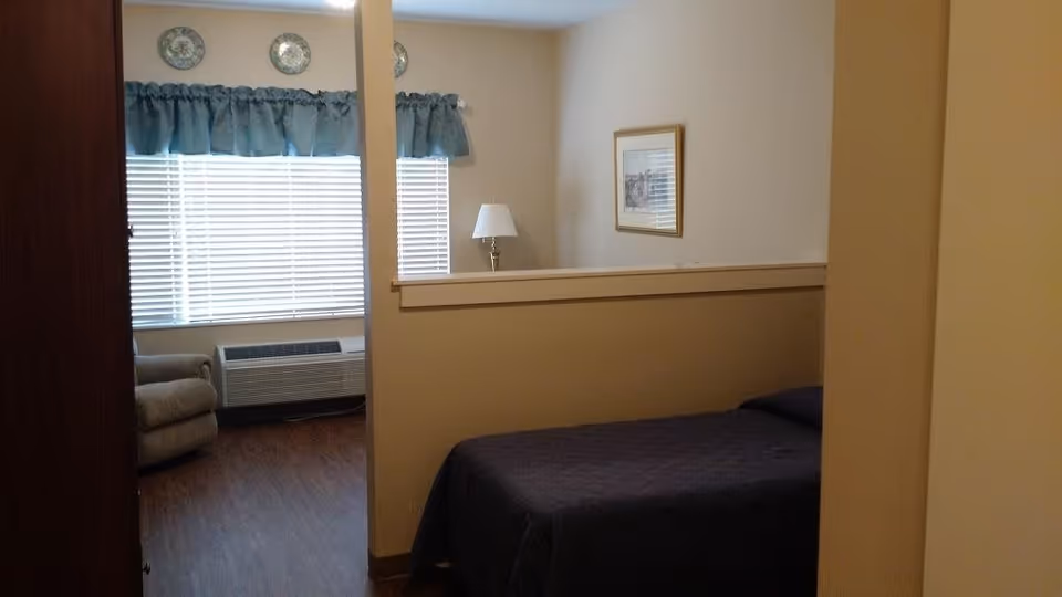 Interior view of a senior living facility room at Alamance House showing a bed with a dark bedspread, a partial wall divider, a framed picture on the wall, a lamp on a small table, a window with blue curtains, an air conditioning unit below the window, and a beige armchair in the corner.
