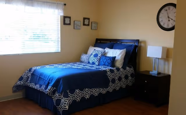 A bedroom with a neatly made bed featuring blue and white patterned bedding and multiple pillows. There is a window with white blinds and a sheer curtain on the left wall. On the right side of the bed is a dark wooden nightstand with a modern lamp. A large round clock hangs on the wall above the nightstand. The room has light-colored walls and wooden flooring.