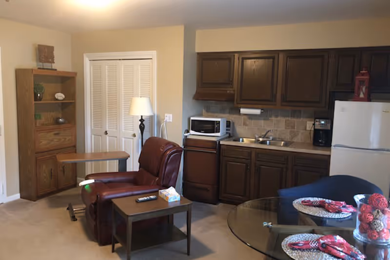 Interior view of a small living space featuring a brown leather recliner chair with a wooden side table holding a remote control and tissue box. Behind the chair is a wooden bookshelf and a floor lamp. The kitchenette area includes dark wooden cabinets, a microwave, a small stove, a sink, a coffee maker, and a white refrigerator. In the foreground, there is a glass dining table set with placemats and decorative items.