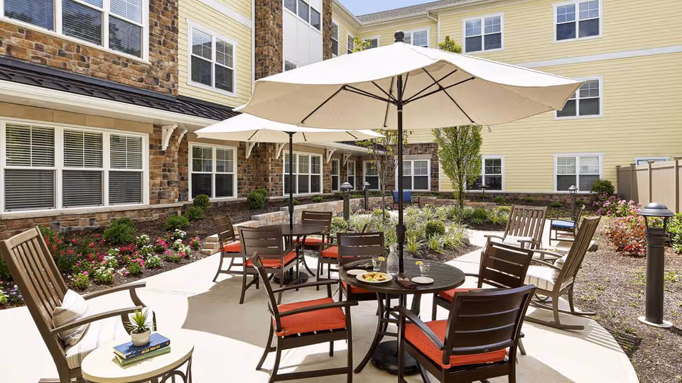 Outdoor patio area at Tappan assisted living nursing home with round tables and chairs featuring red cushions, large white umbrellas providing shade, surrounded by landscaped garden beds and the exterior walls of the building with multiple windows.